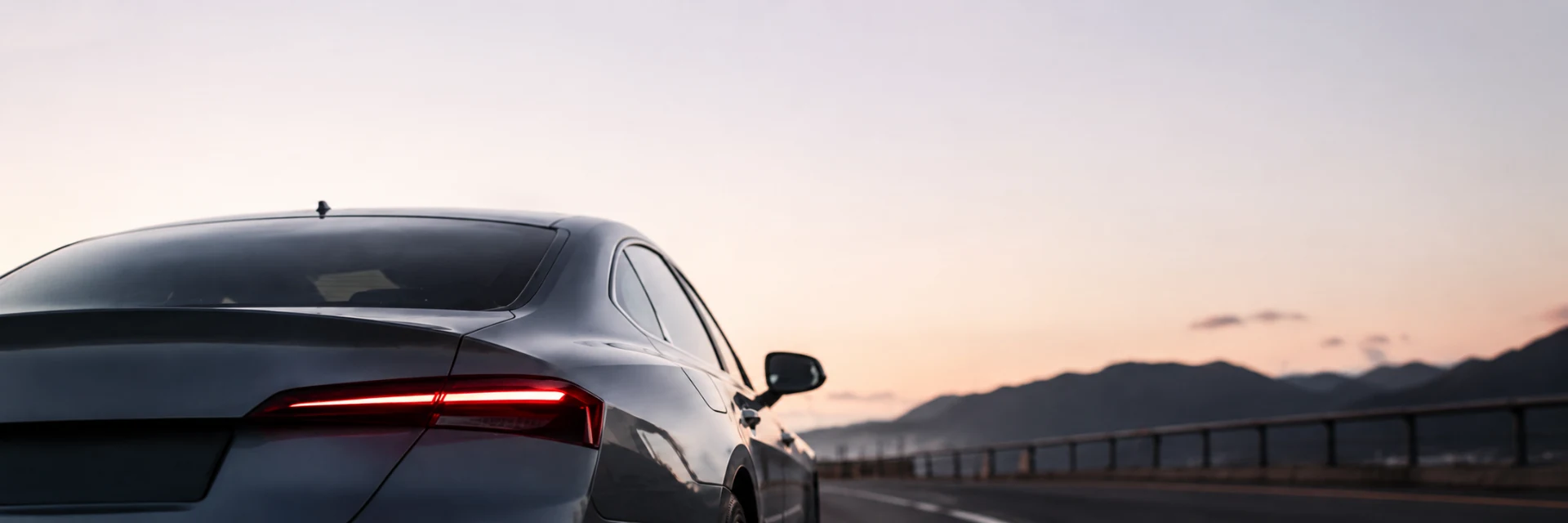 Rear three-quarter view of a grey saloon on an open coastal road at dusk